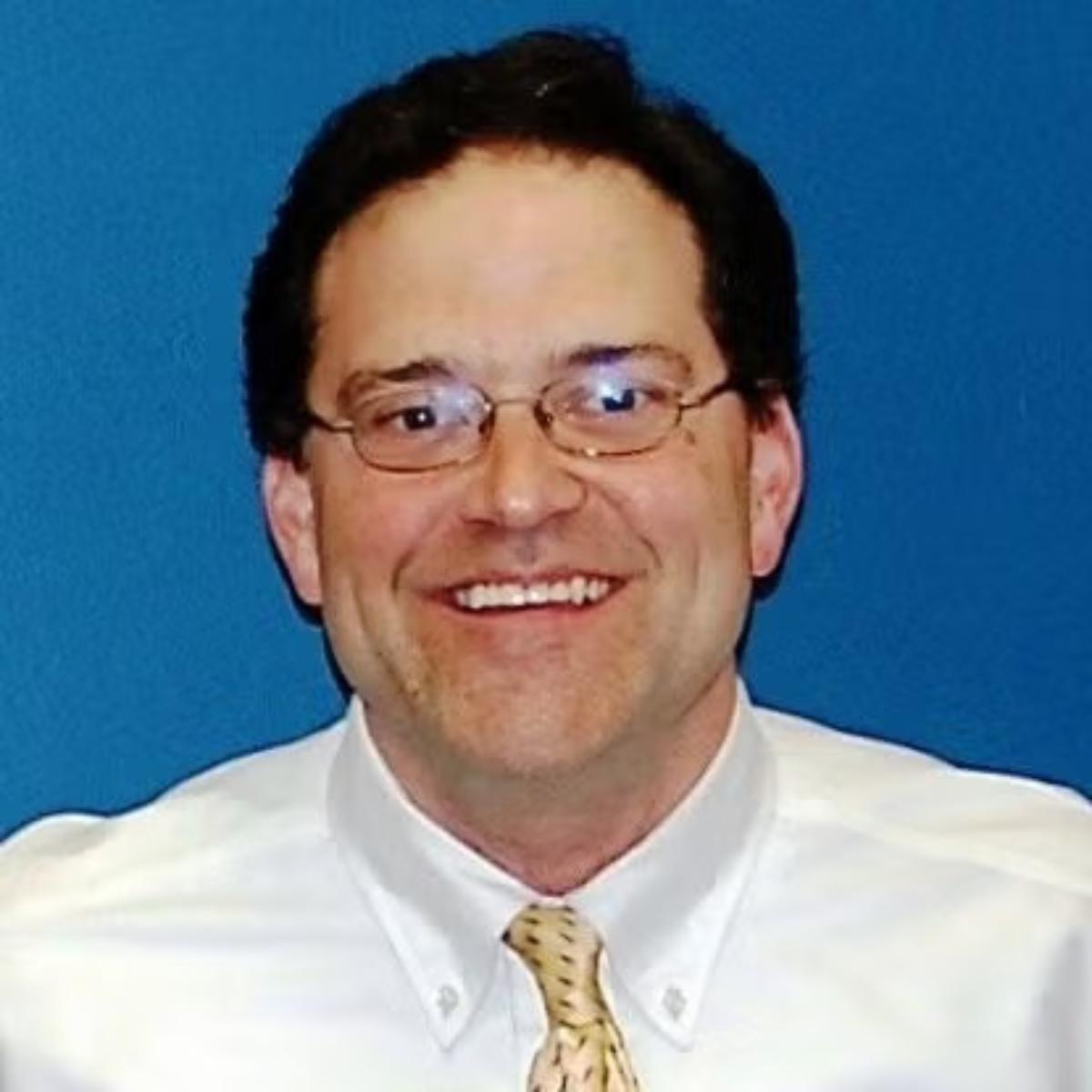Headshot of a smiling man wearing glasses, a white dress shirt, and a patterned tie against a blue background.
