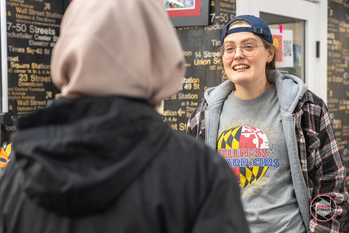 White person with backwards hat and a grey tshirt and plain shirt speaks to a student with a beige head covering and black hoodie, seen from behind, with barbershop wall in background