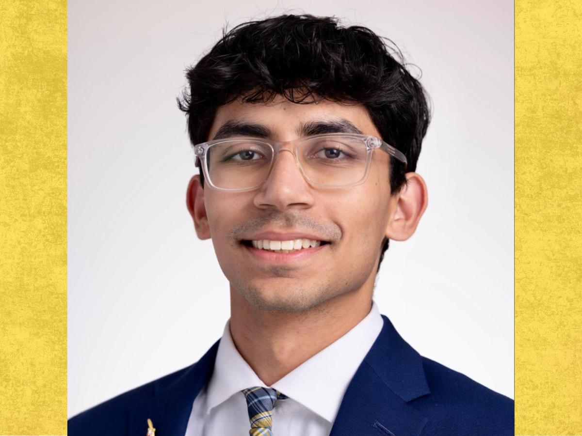 Student wearing dark blue suit jacket, white shirt and colour tie, brown wavy short hair, with glasses on smiles to the camera