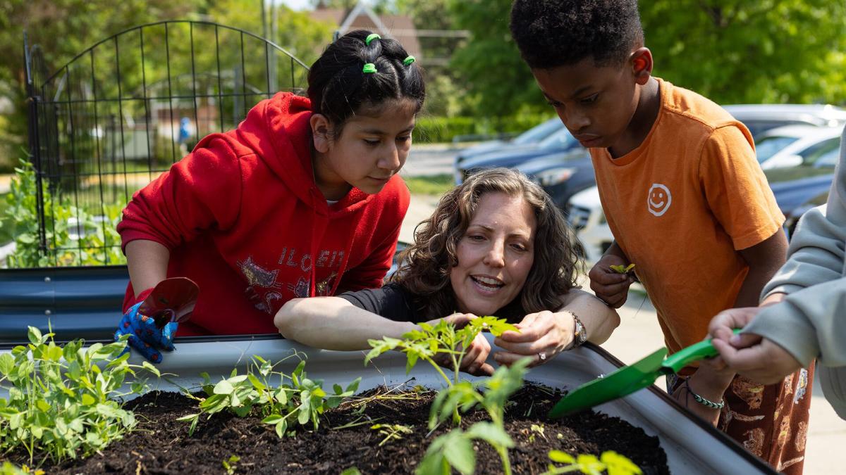 White woman squats down by a raised vegetable bed, two children look over her shoulder at seedling plants