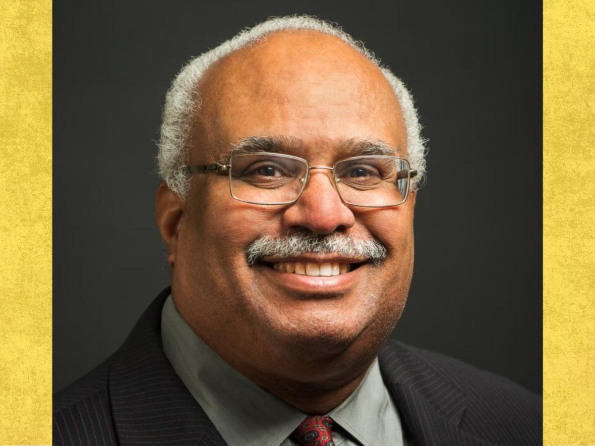 Black older man with greying moustache and rimless spectacle smiles to camera, wearing a dark suit jacket and reddish tie