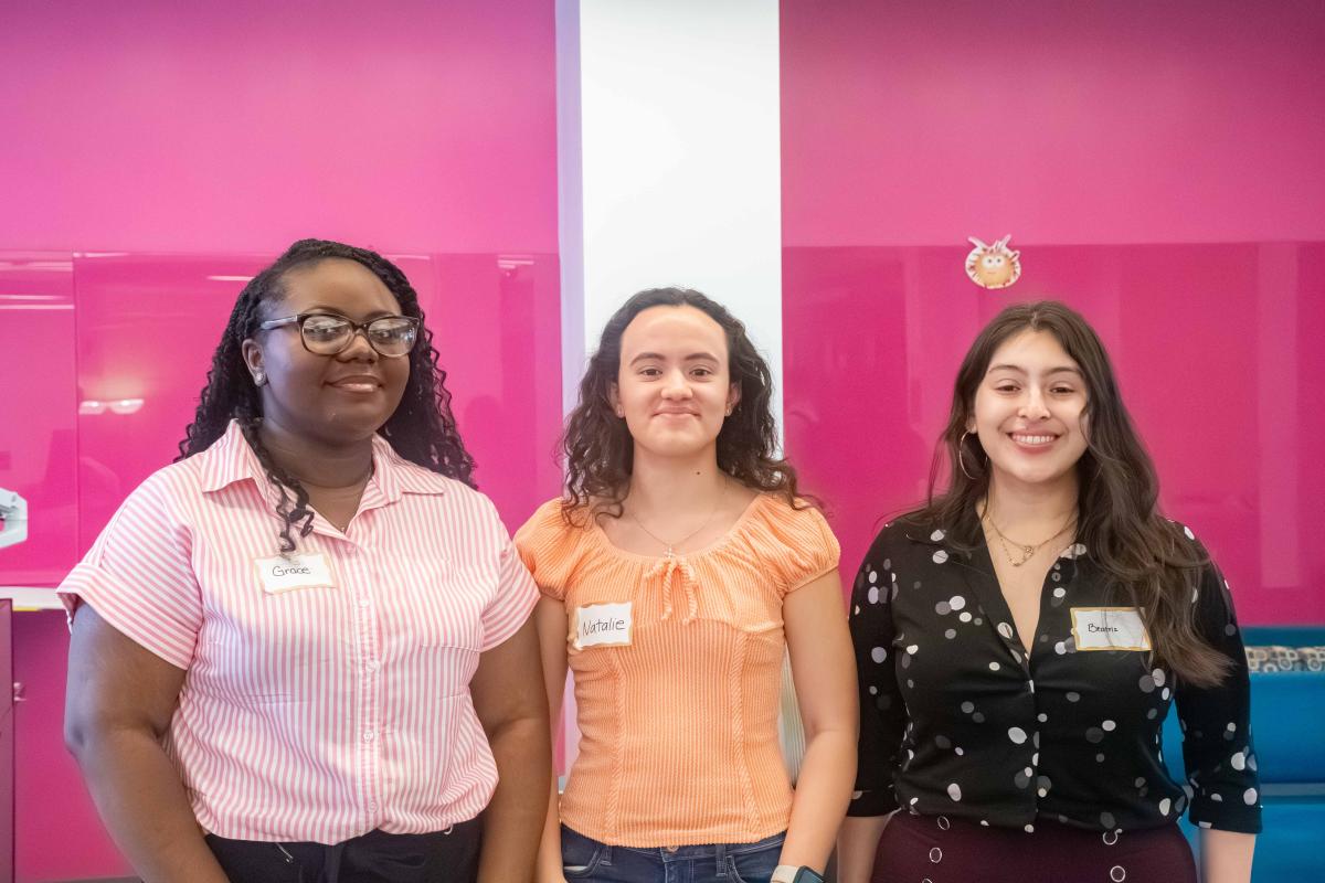 Three female students wearing pink shirt, orange top, black shirt respectively smile to camera in front of a pink background
