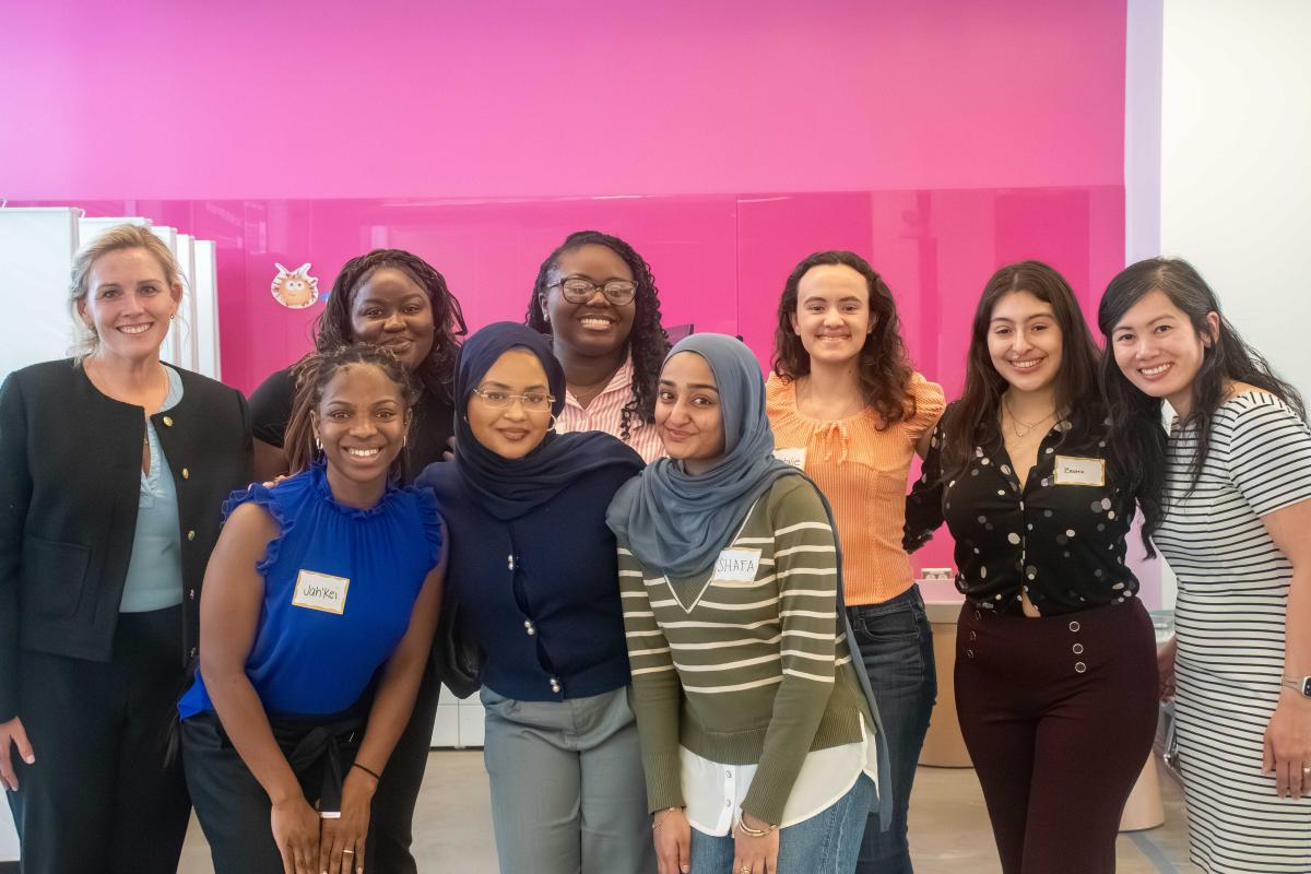 group of several female students and two professors posing smiling in front of pink wall