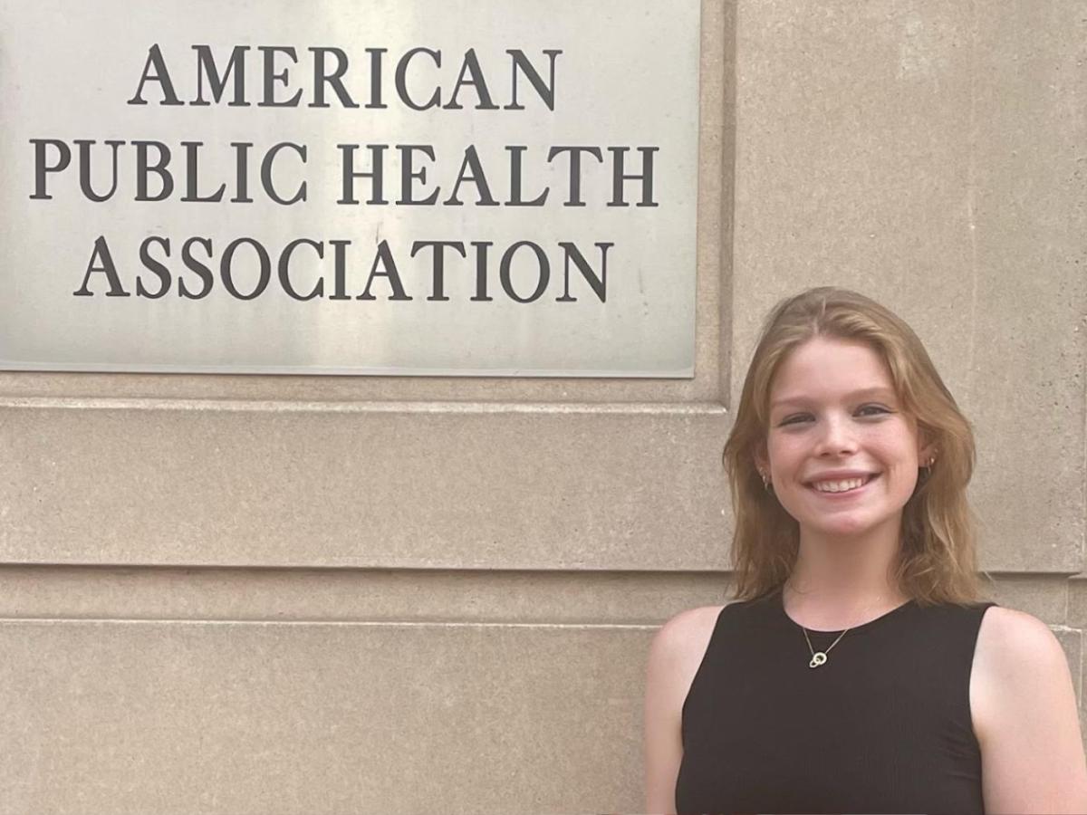 Young white woman in black tank top smiles to camera next to sign for the American Public Health Association