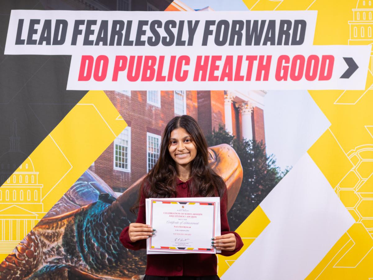 Wearing a dark red top, student with long brown hair smiles to camera, standing in front of SPH banner and holding her award certificate