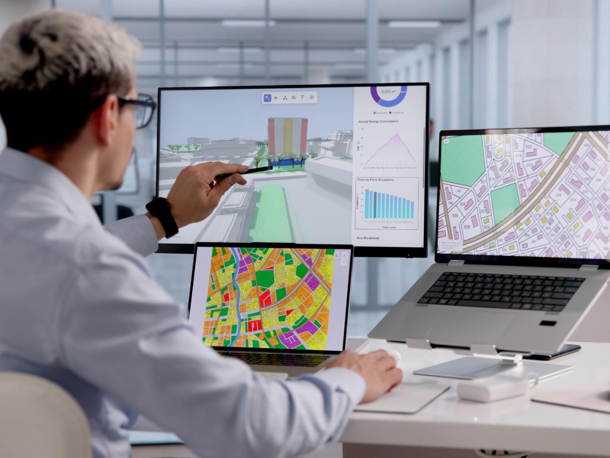 Person in white shirt sits in front of 3 computer screens with different data and maps on them, in office setting (stock photo)