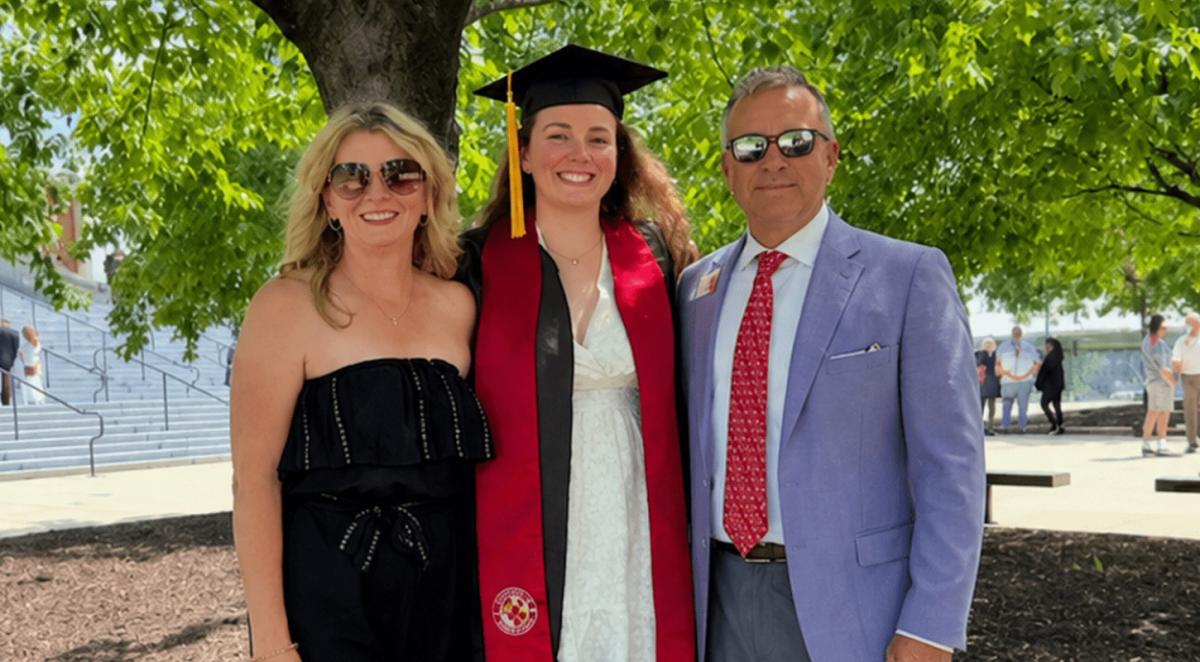 white mother with blond hair in black dress and father in suit with red tie pose in front of tree with white young woman wearing white dress and graduate cap and gown, all smiling