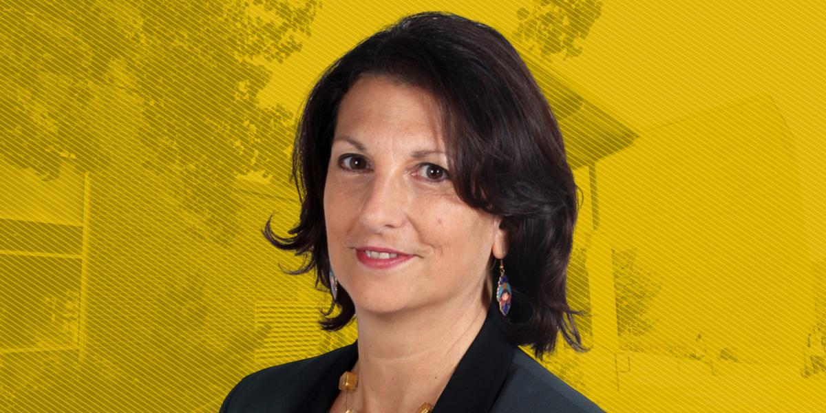 White woman with medium length brown hair, in dark grey suit smiles, with yellow-tinted graphic background of School of Public Health building and some tree foliage