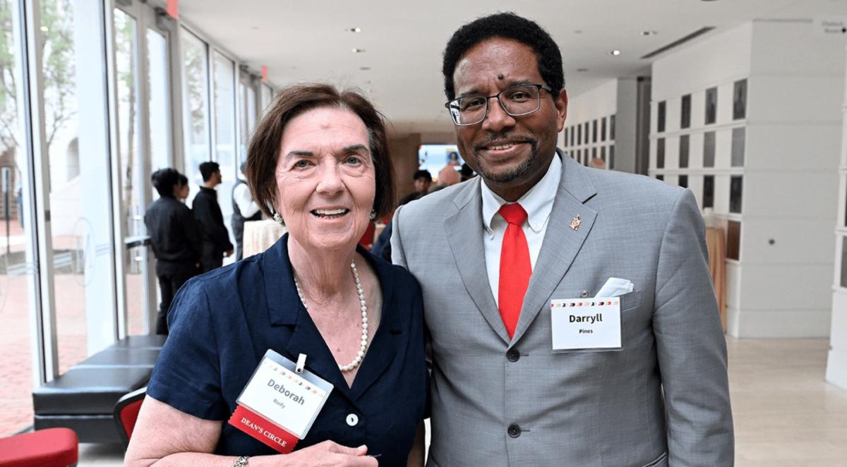 Older white woman in navy suit dress poses with older Black man - university president - wearing glasses and agrey suit with red tie, both small smiling