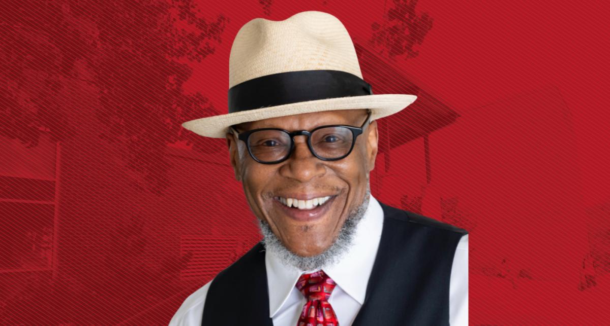 Black man in glasses with a white hat with black ribbon and vest, white shirt and red tie, smiling — background is a red tinted graphic of SPH building