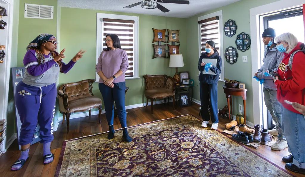 Black woman in glasses and purple tracksuit and headscarf gestures to a group of four people standing in a room with intricate carpet, light green walls and art items on the walls