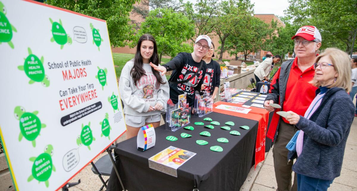 Students, staff and parents in UMD tshirts and hats gather around a table, looking at a big board displaying all the programs at School of Public Health (outdoors, trees in background)
