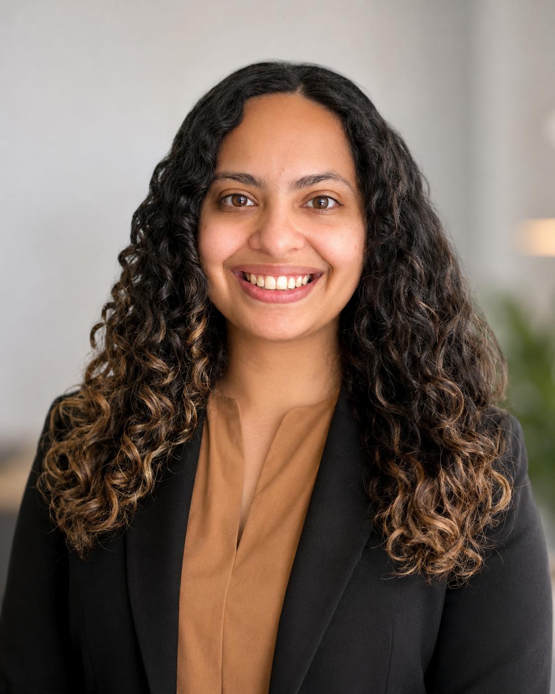Woman with long curly brown hair smiling at camera wearing a black blazer.
