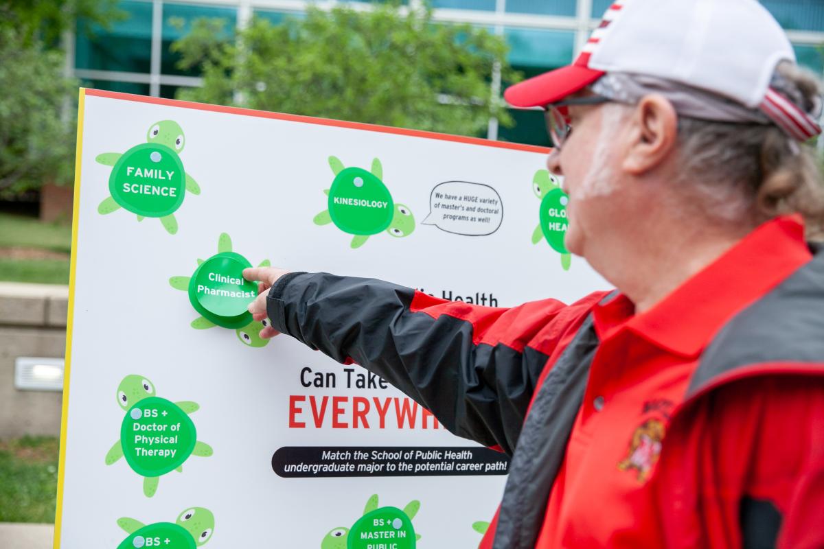 Older white man in UMD shirt, wearing white hat and glasses, pins a green circle that says Clinical Pharmcologist to a board with all the SPH degree programs (matching career to degree)