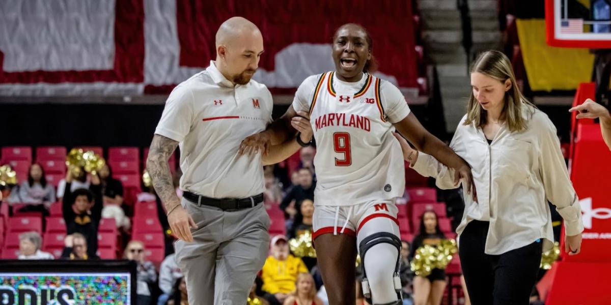 White man in grey slacks and white Maryland shirt helps injured and grimacing Black female basketball player in Maryland kit to walk, assisted by a white woman in black trousers and light button down shirt, on basketball court