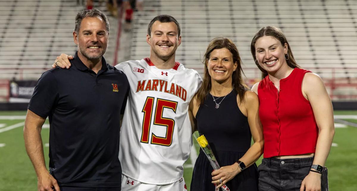 Family poses on lacrosse pitch with two parents, mother holds a flower, father on far left wears black Maryland polo, son to his right wearing Maryland lacrosse kit and daughter on far right wearing a red top and jeans, all smiling