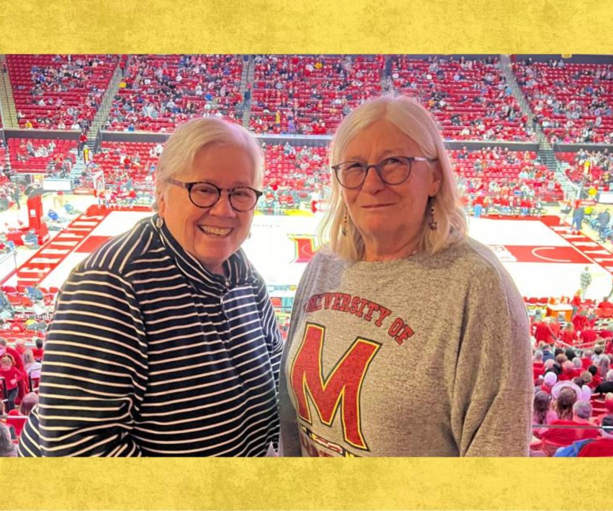 Two women smile for a photo inside a University of Maryland basketball arena with the court and spectators in the background.