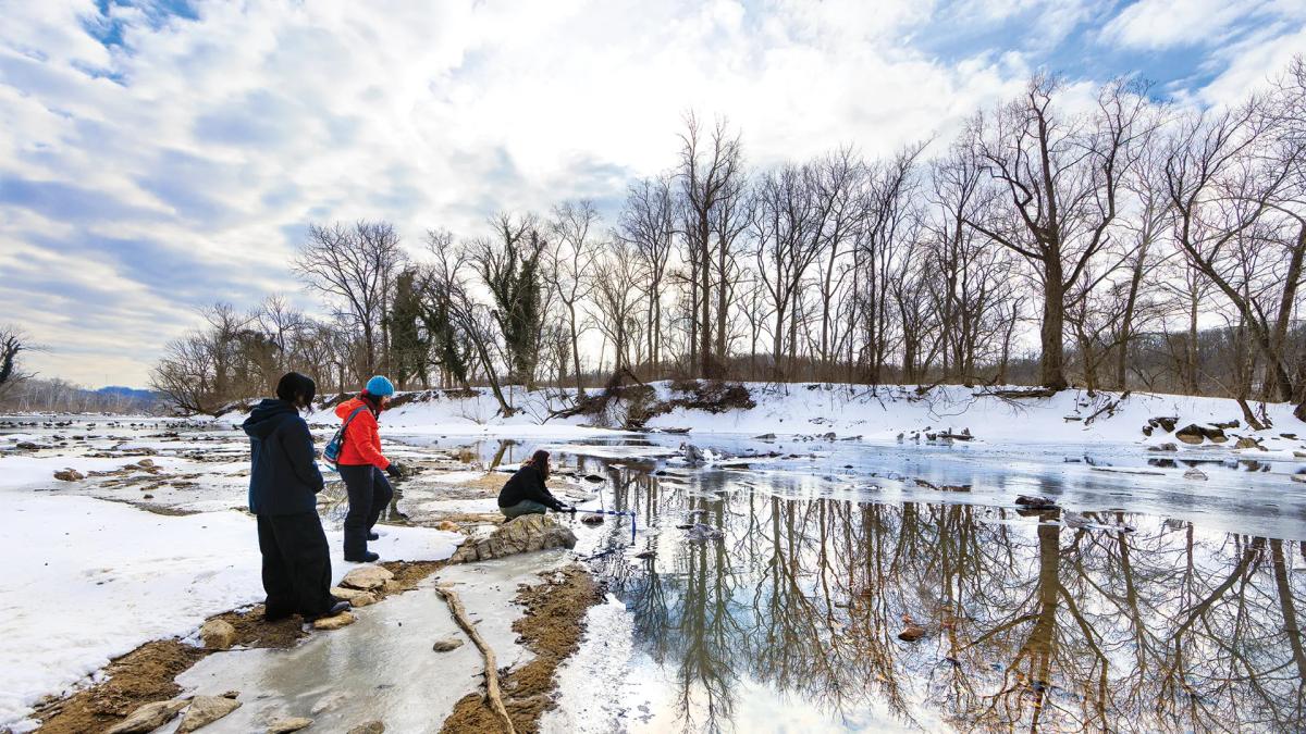 wide angle of three people by a snowy river scene with trees in background, bright sky, and one person kneeling down by the water to take a sample with a bottle. 