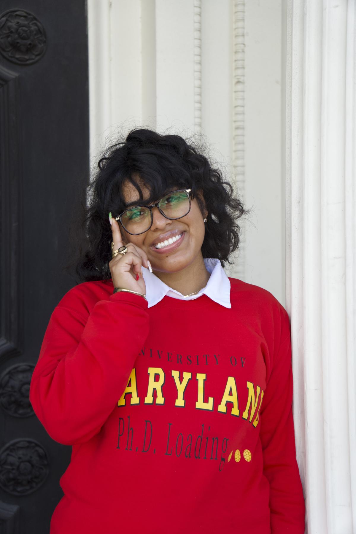 Woman facing camera touching glasses smiling with a Maryland shirt on.