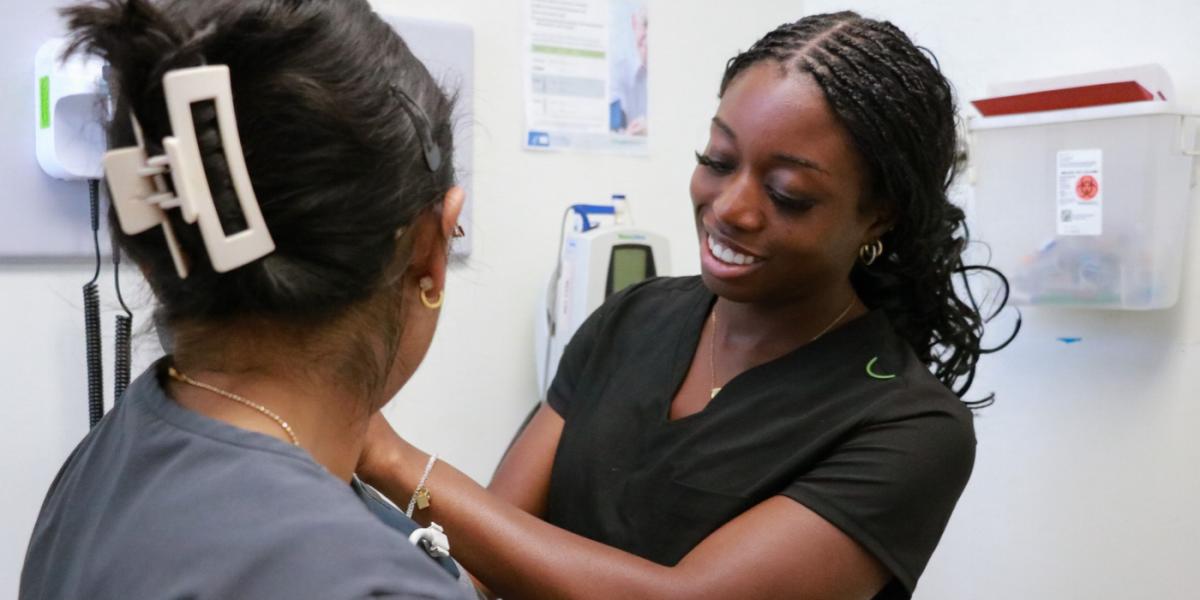 A black woman with braided hair in black v-neck scrubs tends to a woman with her back to us, long brown hair up in a white hairclip, wearing a grey top, background looks like a clinic room