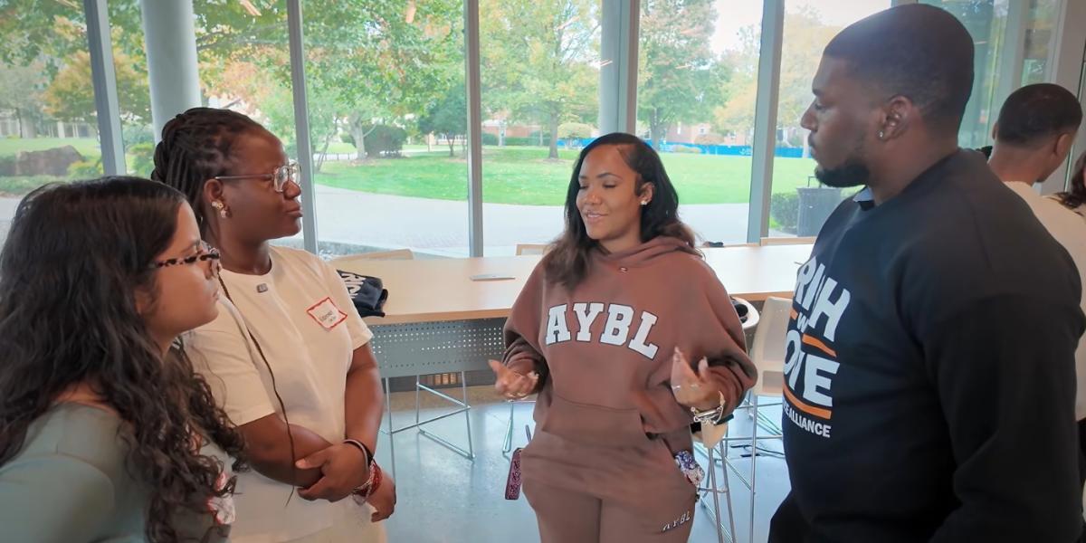 Four students of colour stand together, three listen to one student speaking, who wears beige sweatshirt and pants, in a room with floor to ceiling windows, trees, grass and pathways are seen in background