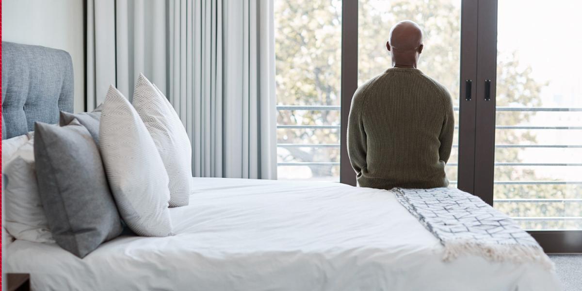 Black man sits at the edge of a bed looking out the window, with back to viewer