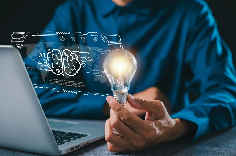 Photo illlustration of person holding lightbulb while sitting in front of computer with scan of brain and term "AI" in front of the computer.