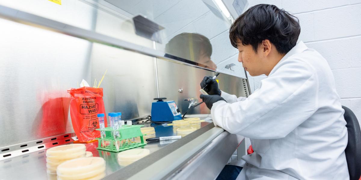 Asian man with dark hair in a white lab coat tests water samples in a lab environment, with petri dishes in the foreground on the counter