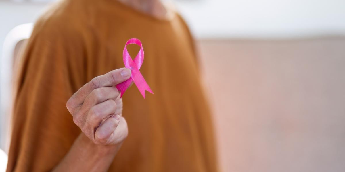 An older woman, only shown from neck down to waist, wearing a mustard coloured tshirt, holds a pink ribbon (for breast cancer awareness) up towards the camera