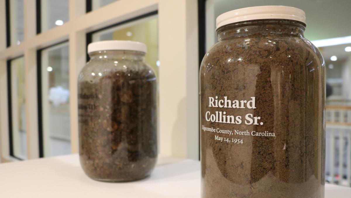 Two jars of soil on a white counter with names, dates and places written on the bottles, windows in background