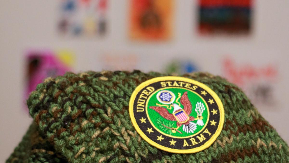 A knitted green fabric with a patch of the United States Army, in background some artworks on the wall. 