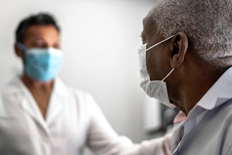 Older African American man seated in doctor's office with health care professional standing up, both are wearing masks.