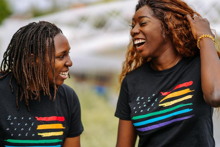 A Black queer couple laughing and looking at each other in t-shirts with rainbow stripes that say Love. 