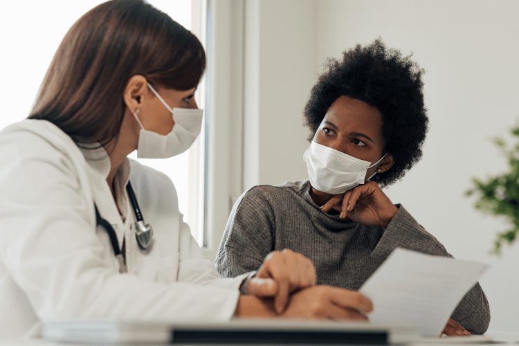 A white female doctor explaining a diagnosis or health information to an African American woman patient, both wearing masks