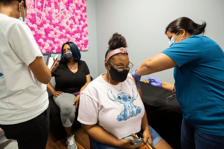 A young Black woman receives a COVID vaccine at a Black beauty salon with other women sitting nearby