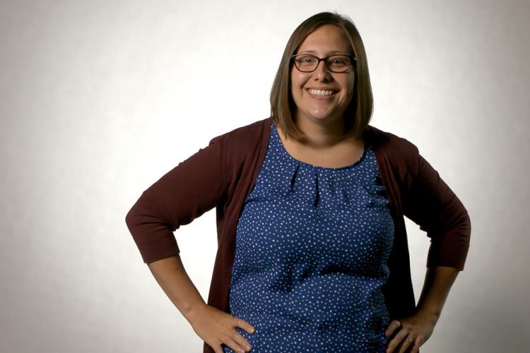 Jessica Fish, a white cis gendered woman with shoulder length brown hair wearing glasses, stands with her hands on her hips