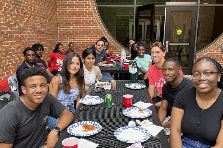 A group of African American, Latino and white UMD college students sit outside at a picnic table with pizza on paper plates. 