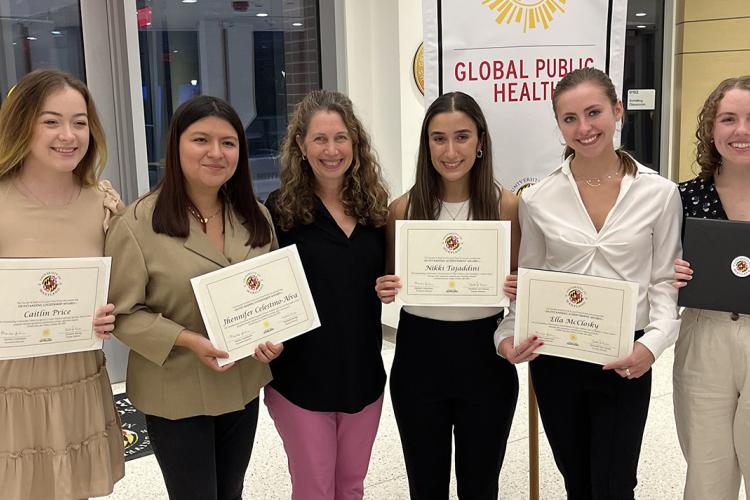 A group of five college students hold awards certificates, and a faculty member stands in the middle of the group smiling.