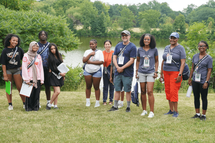 Students and adults stand together smiling to camera in green park area in front of river