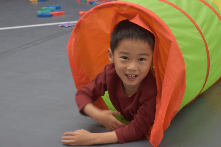 young asian boy in red longsleeve shirt smiles, as he crawls out of a mesh tunnel in the gym