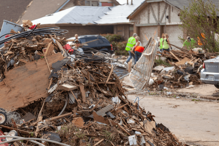 debris of destroyed house is piled up to the left, in background people in neon safety vests clean up