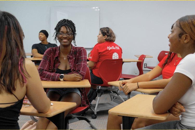 students sit at single desks in a small group talking in a classroom space, with another student group visible in the background