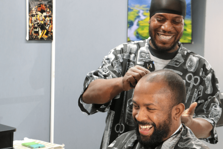 Black man barber makes a joke while his Black man customer laughs, sitting in chair