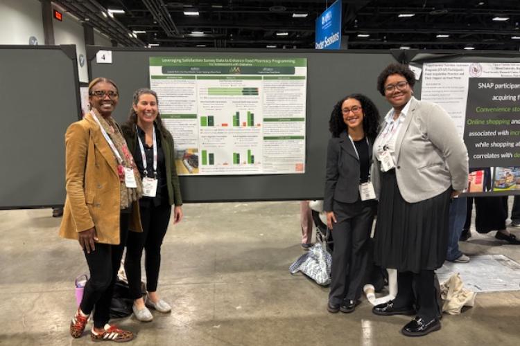 White woman masters student stands with three other women students, smiling to camera in front of a public health poster at a conference