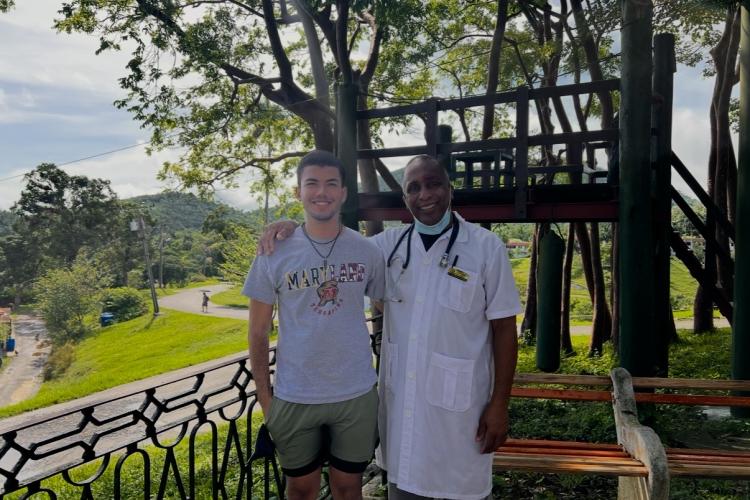 Student and professor pose in front of a bench and tree at a rural community-based healthcare facility in Cuba