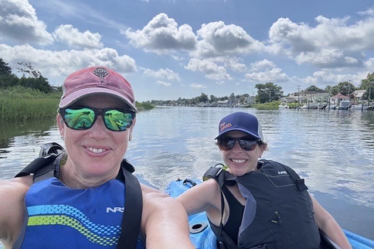 two white women in hats and sunglasses smile sitting in kayaks on the river