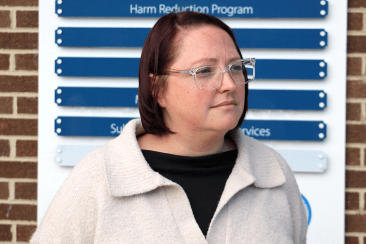 White woman in glasses and a white v-neck sweater stands in front of HEALTH HUB sign, looking off to the right