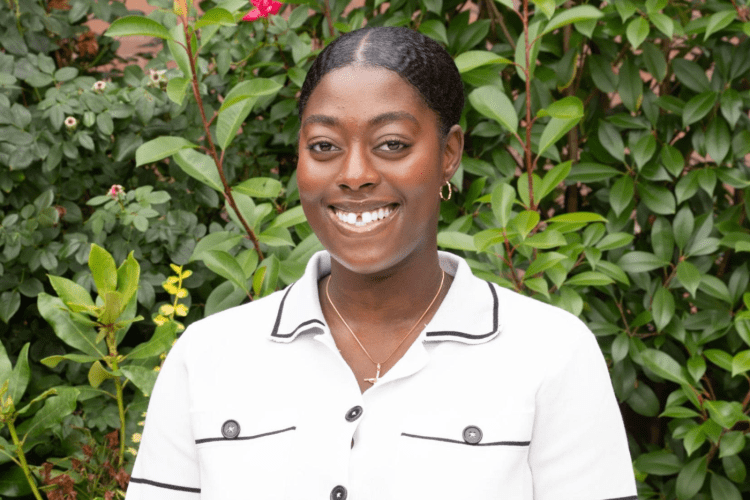 African American student smiles to camera, wearing white shirt in front of green bushes. 