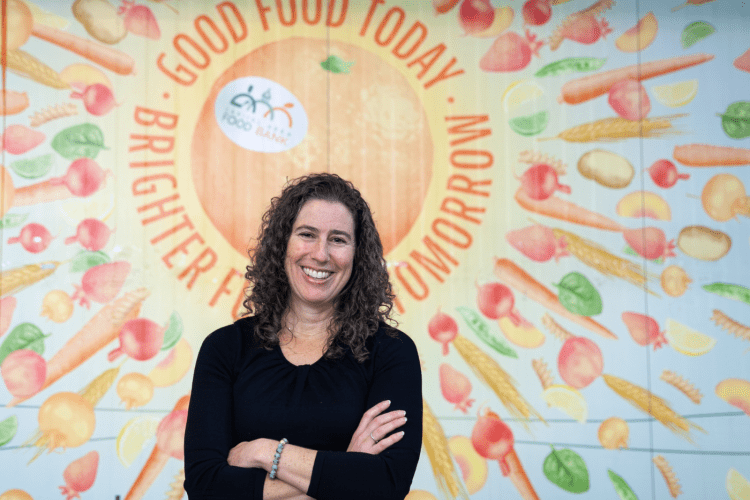 White woman with curly long hair in a black top smiles in front of a wall mural with vegetables saying GOOD FOOD TODAY