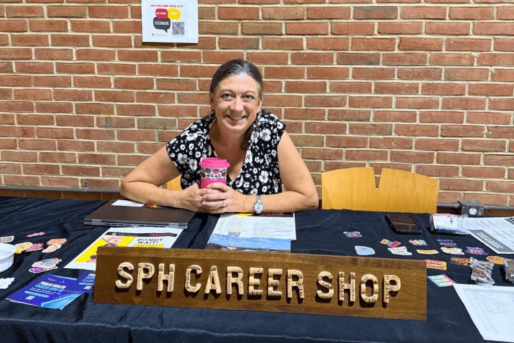white woman sits at a table with sign for SPH Career Shop, wearing black and white floral shirt, smiling at camera